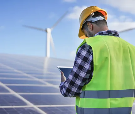 a man wearing a hard hat and a yellow vest wearing a chequered long sleeve shirt looking at his table and solar panels and wind turbines in the distance with a blue sky background